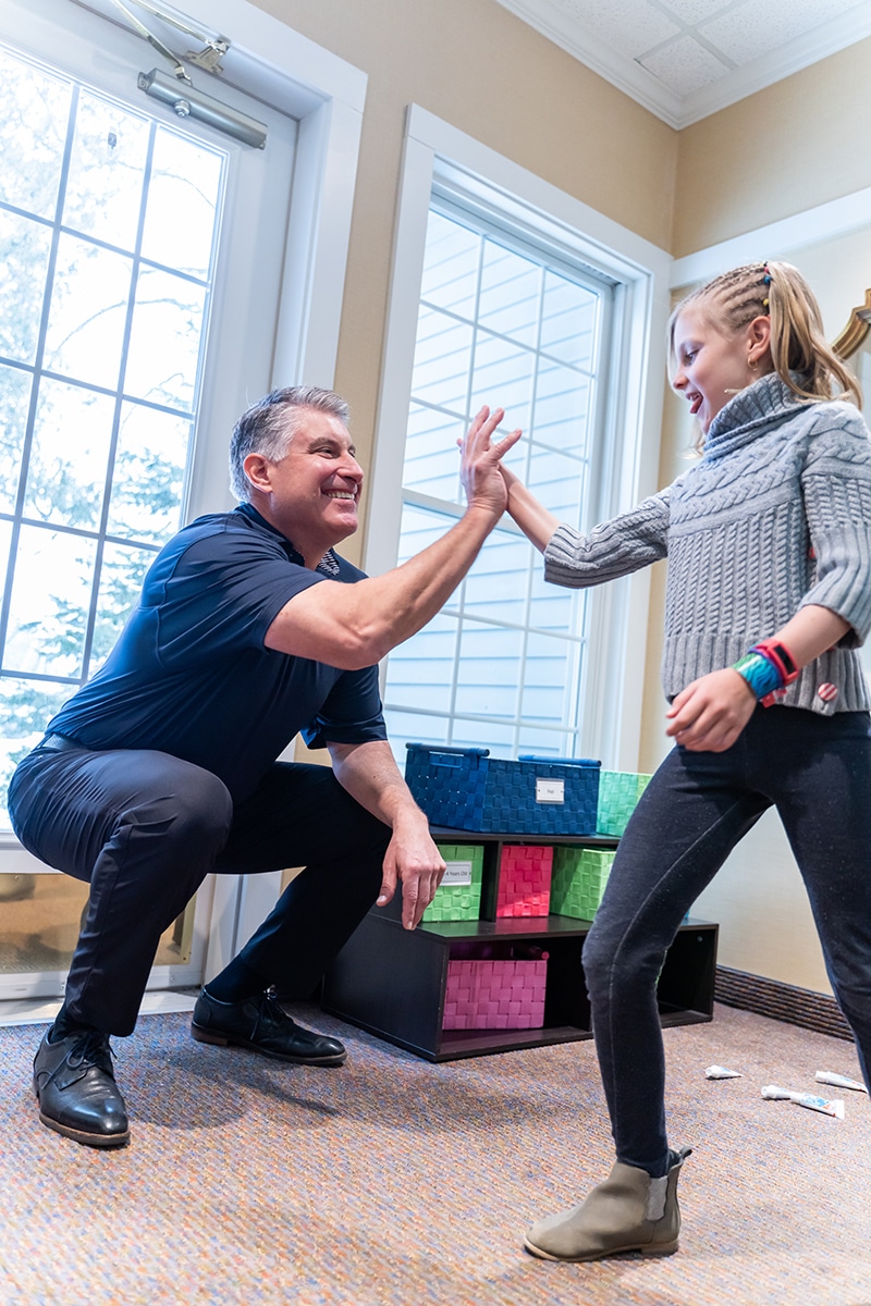 Adult and child high-fiving in bright dental office with large windows and colorful toy blocks.