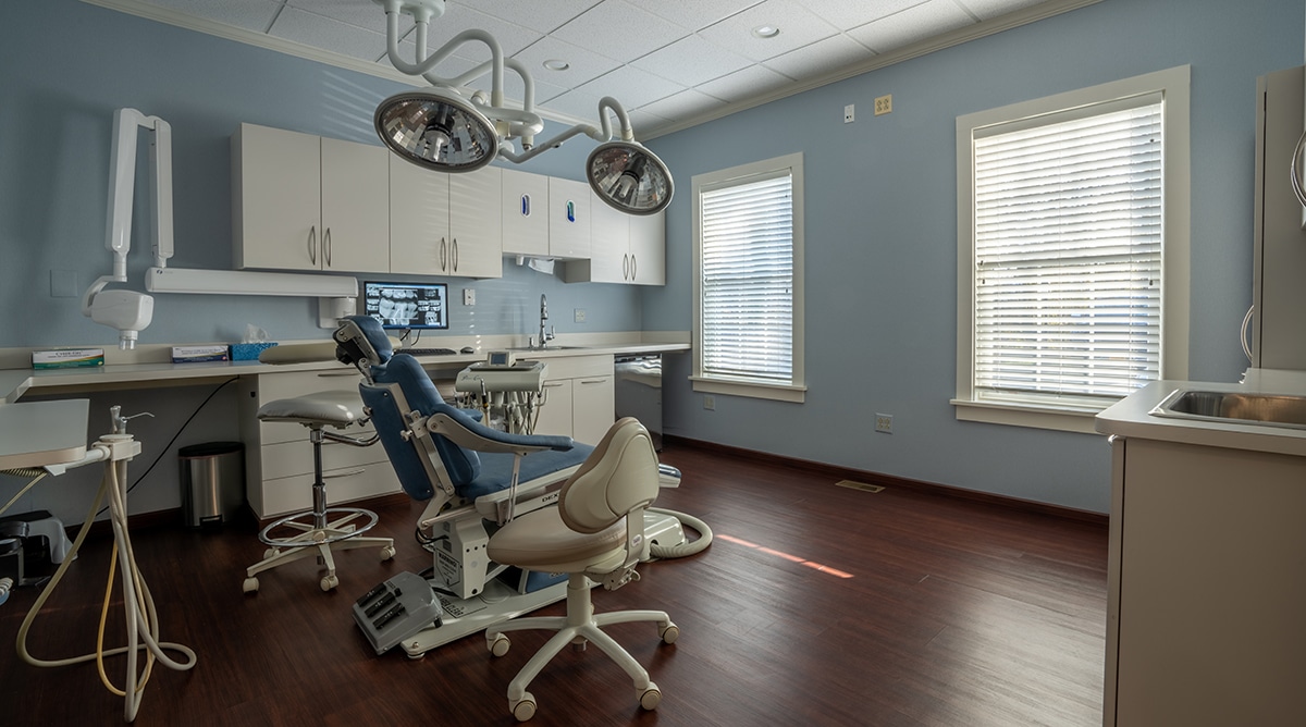 Modern dental operatory with blue patient chair, overhead lights, white cabinetry, and clinical equipment.