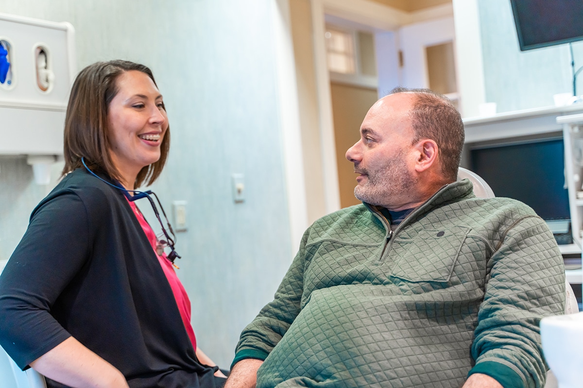 Female dental staff member smiling while speaking with male patient in modern dental office setting.