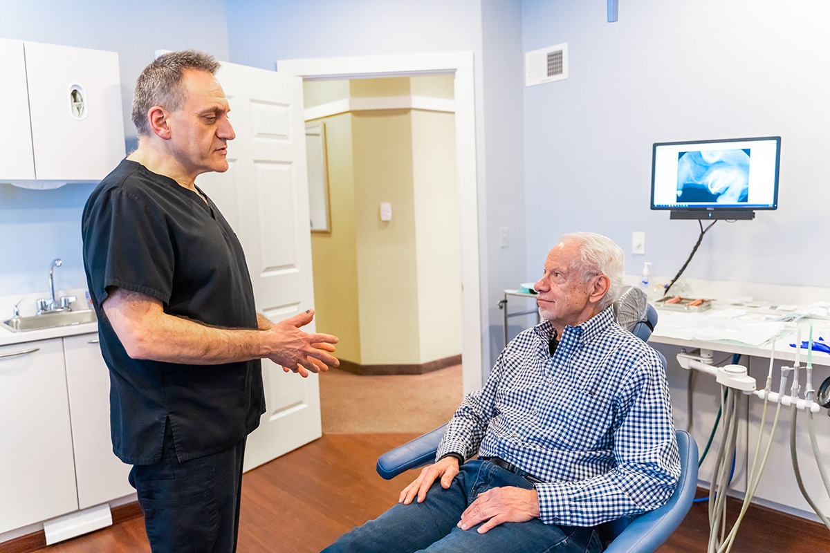 Dentist speaking with senior patient in dental exam room with X-ray on monitor.