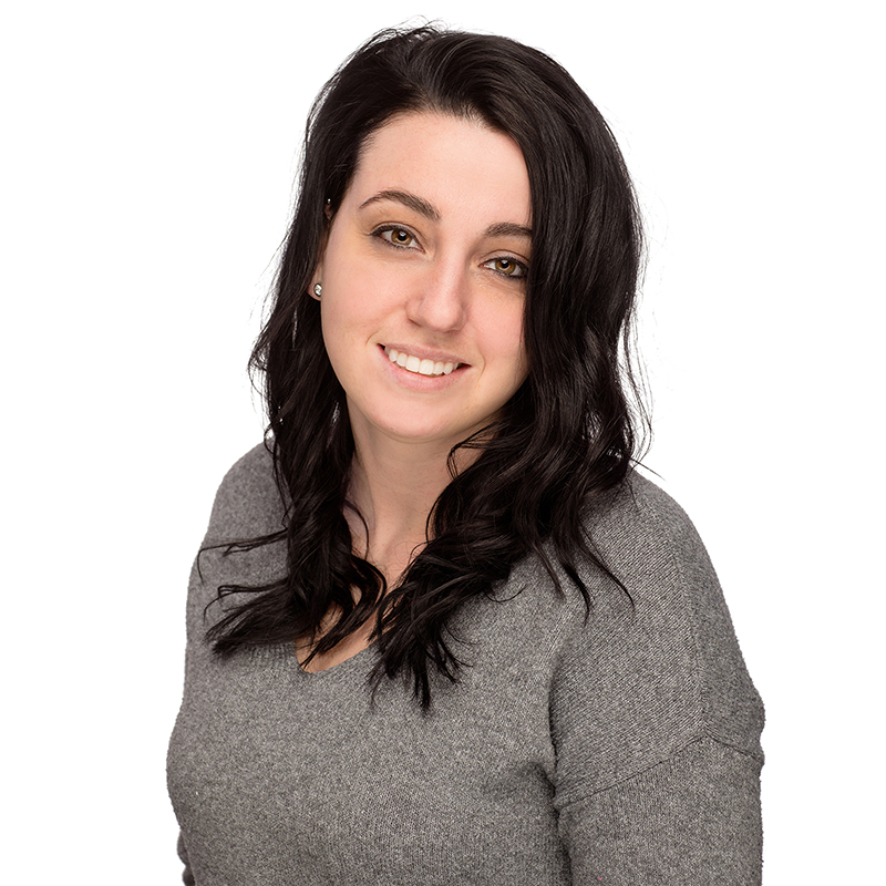 Professional headshot of smiling woman with dark hair wearing gray sweater against white background.