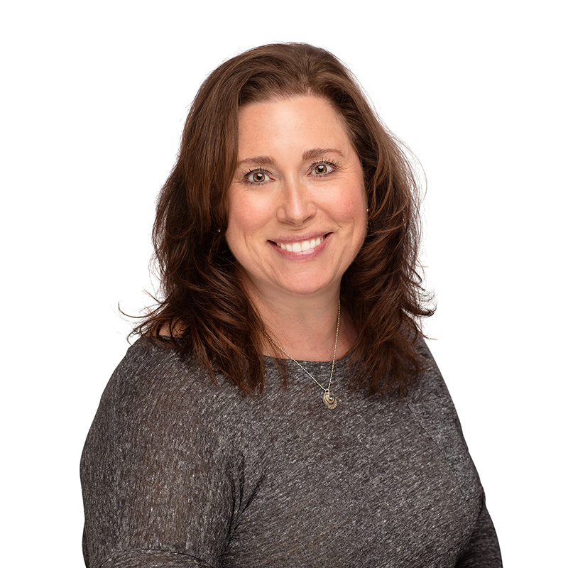 Professional headshot of smiling woman wearing gray top against white background.