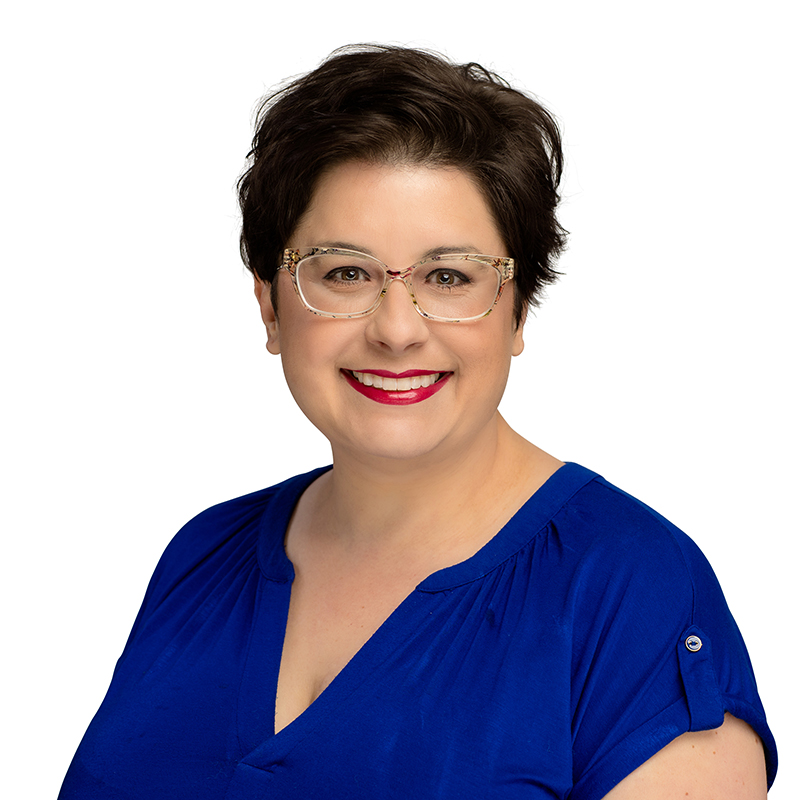 Professional headshot of woman with short dark hair, glasses, and blue scrubs against white background.