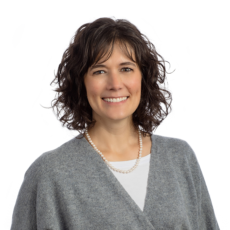 Professional headshot of smiling woman wearing gray cardigan and pearl necklace against white background.