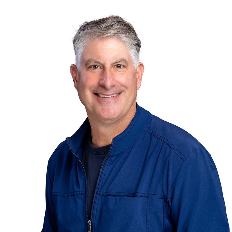 Professional headshot of smiling man in blue jacket against white background at dental office.