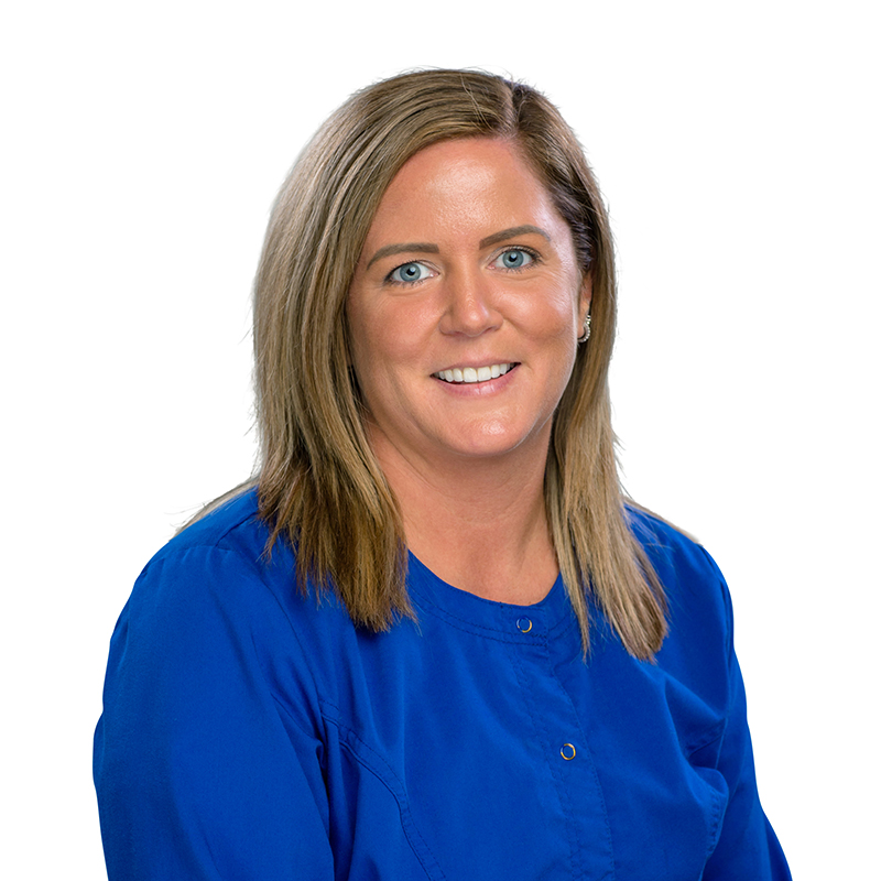 Professional headshot of woman in blue scrubs smiling at camera, dental office setting.