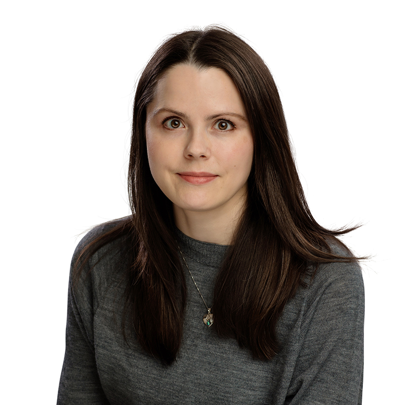 Professional headshot of woman with long dark hair wearing gray sweater against white background.