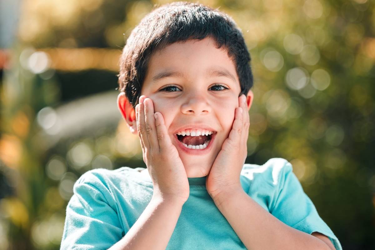 Young boy in turquoise shirt smiling with hands to face outdoors in sunny garden setting.