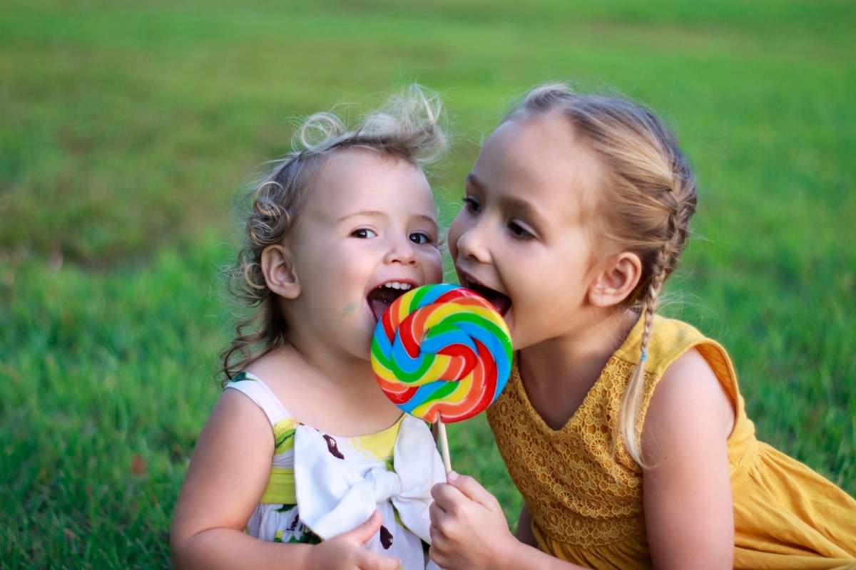 Two young girls sharing a colorful lollipop outdoors on green grass background.