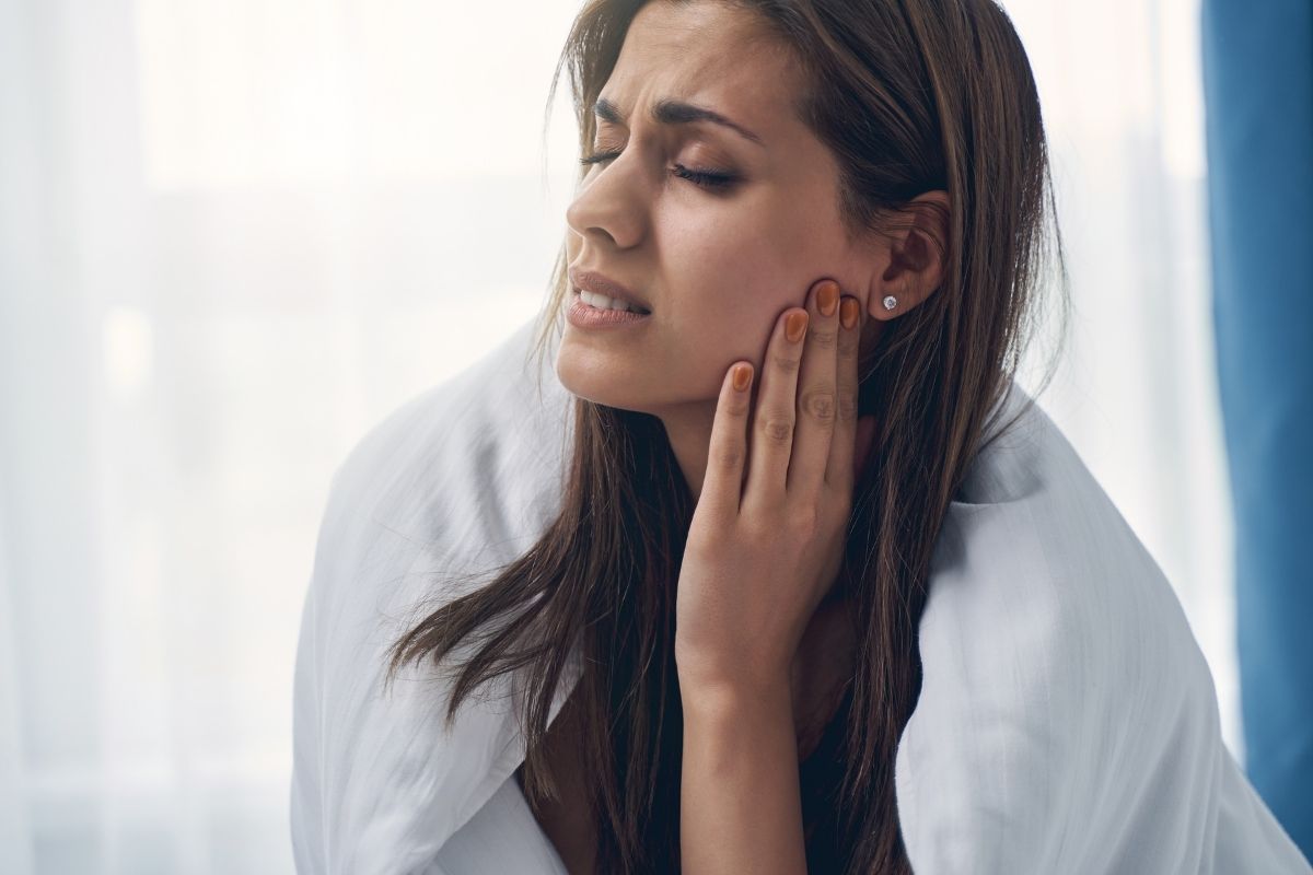 Woman experiencing jaw or tooth pain, holding her cheek in discomfort against bright window background.